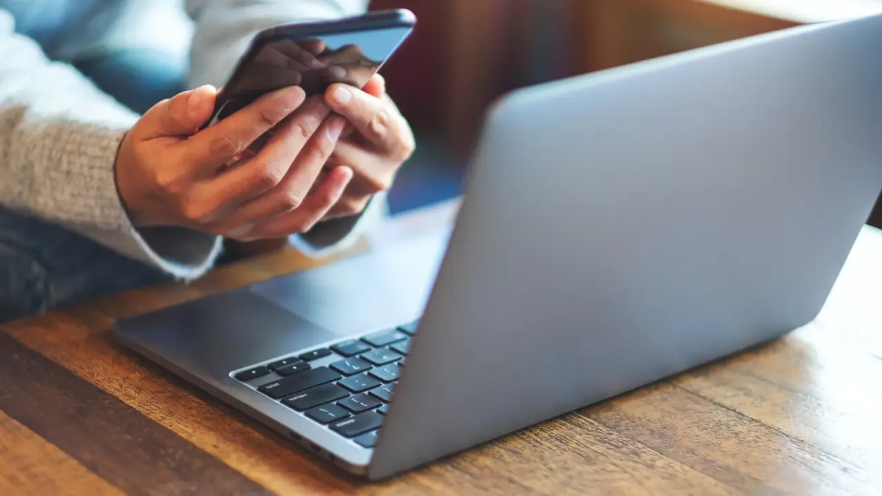 Nahaufnahme von Händen einer Frau, die gerade am Laptop sitzt und im Handy tippt